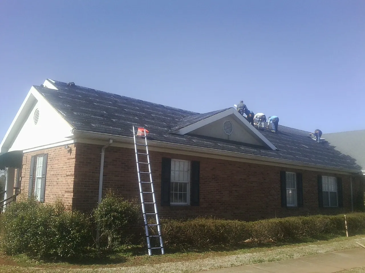 Crew tearing shingles off a brick ranch roof with a ladder set — Emergency Roof Repair work by our crew in Mount Horeb