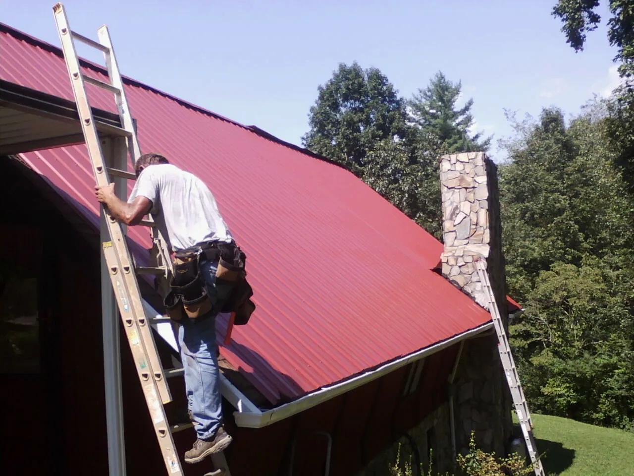Roofer on a ladder beside a red metal roof and stone chimney — Soffit & Fascia Repair by Roofing Brothers in Mount Horeb